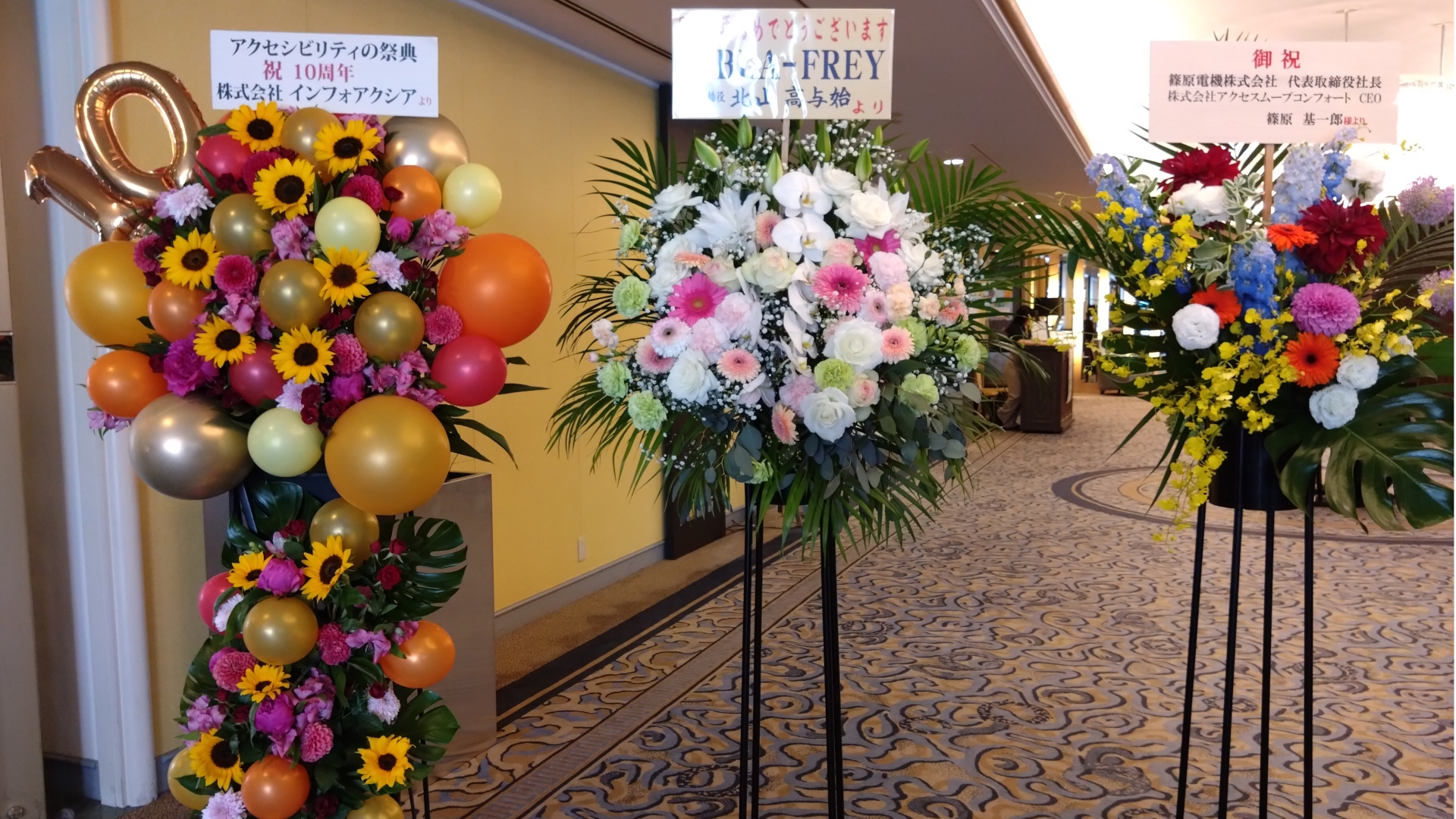 写真：イベント会場の入口。10周年のお祝いの、3つのスタンド花。色とりどりの花や丸い風船、10を表す金色の風船が飾られている。贈り主は、株式会社インフォアクシア、BLA-FREY、篠原電機株式会社・株式会社アクセスムーブコンフォート。